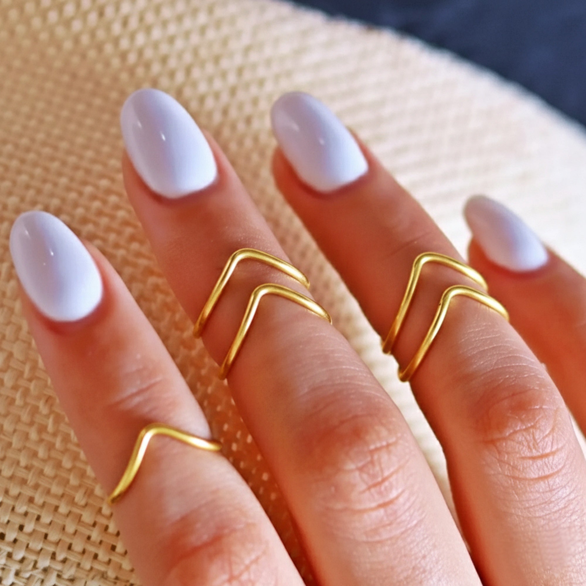 Close-up of a hand wearing three gold rings with white nail polish on a textured beige background